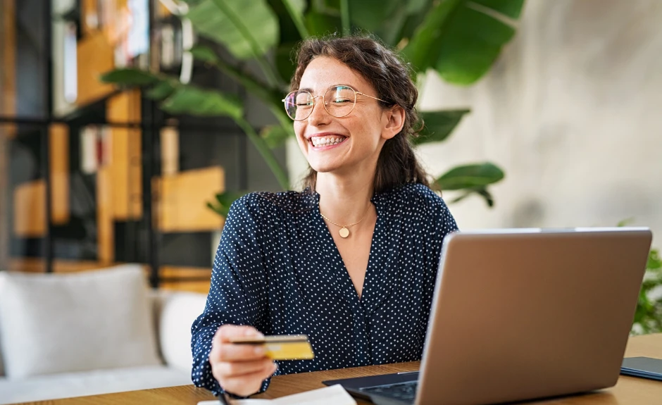 Mujer feliz trabajando en el ordenador y sosteniendo una tarjeta TF Mastercard Oro.
